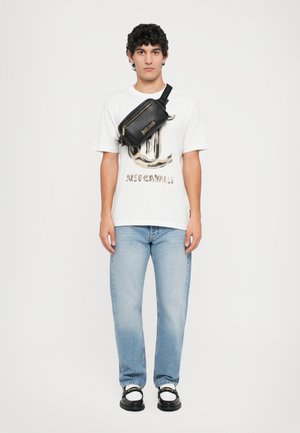 Zwarte schoudertas met rits en gouden accenten, witte T-shirt met korte mouwen en grafische print, lichtblauwe spijkerbroek en zwarte sneakers.