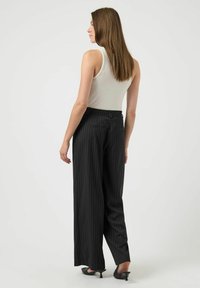 Vrouw met lang bruin haar, gekleed in een witte mouwloze top, een zwarte pinstripe wijde broek en zwarte hoge hakken, kijkt naar links.