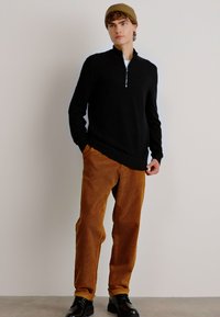 Black knit sweater with zip collar, paired with tan corduroy pants. Model wears black shoes and a brown beanie, standing against a neutral background.