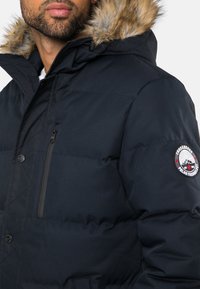 Marineblauer Puffer-Jacke mit einer Kapuze mit Fellbesatz, mit Reißverschlusstaschen, gestepptem Design und Logo-Patch am Ärmel. Strukturierter Stoff für Wärme.