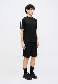 Fekete sportos póló fehér csíkokkal az ujjain, texturált fekete rövidnadrággal, amely geometriai mintával díszített, fekete zoknival és cipővel párosítva.