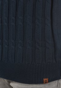 Pull en maille bleu marine avec des motifs de torsades, des poignets et un ourlet côtelés, avec un petit patch logo marron en bas.