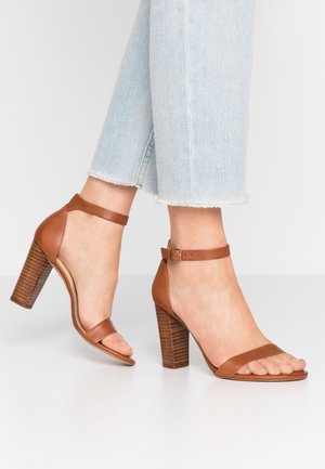 Sandales à talons blocs en cuir marron avec bride à la cheville, portées avec un jean court à bords effilochés bleu clair, sur un sol blanc.