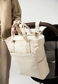 Beżowa torba płócienna z podwójnymi uchwytami u góry, z przednią kieszenią i regulowanymi paskami, przymocowana do uchwytu wózka.