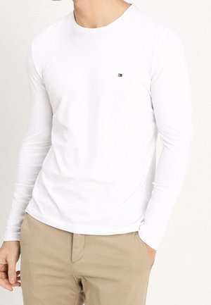 Weißes Langarm-T-Shirt mit rundem Ausschnitt und kleinem Logo-Akzent. Hergestellt aus leichtem Stoff, figurbetonter Schnitt, kombiniert mit beigen Hosen.