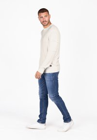 Crème gebreide trui met geribde zoom en manchet, gecombineerd met blauwe slim-fit jeans en witte sneakers. Model staat tegen een witte achtergrond.