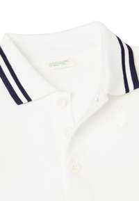 Polo bianco con colletto a righe navy, patta con tre bottoni e logo di un coniglio ricamato sul petto. I dettagli sono sobri e classici.