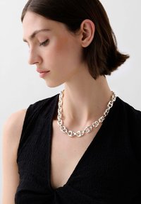 Silberne Halskette mit miteinander verbundenen, länglichen Gliedern, die um den Hals über einem strukturierten schwarzen Oberteil getragen wird. Einfaches und modernes Design.