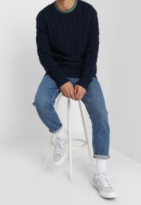 Marinblå kabelstickad tröja med rund hals, i kombination med ljusblå jeans och grå sneakers. Strumporna är vita; sitter på en vit pall mot en ljus bakgrund.