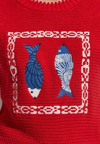 Tissu tricoté rouge avec deux poissons décorés de motifs bleus et blancs brodés à l'intérieur d'une bordure en maillon de chaîne blanche.