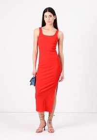 Red sleeveless midi dress with ribbed texture, side slit, and a fitted silhouette. Paired with strappy nude sandals and a blue clutch.