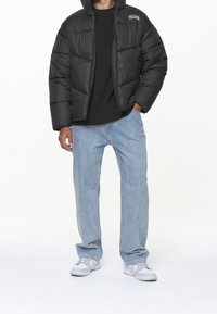 Svart pufferjacka med huva, quiltat mönster, dragkedjestängning, bärs över en svart skjorta, tillsammans med ljusblå jeans och grå sneakers.