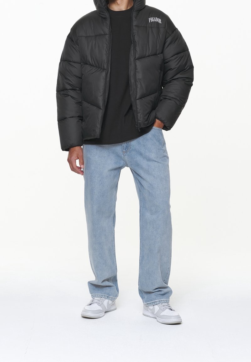 Svart pufferjacka med huva, quiltat mönster, dragkedjestängning, bärs över en svart skjorta, tillsammans med ljusblå jeans och grå sneakers.