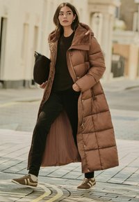 Abrigo largo de plumas marrón con cuello forrado de piel, cierre de cremallera y diseño acolchado, combinado con pantalones negros y zapatillas marrones.