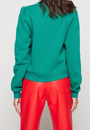 Personne portant un haut vert à manches longues et un pantalon rouge vif taille haute, vue de dos sur un fond clair uni.