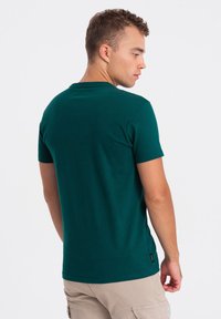 Zelené bavlněné tričko s krátkým rukávem, kulatým výstřihem a volným střihem, které má malou logo aplikaci blízko lemu na levé straně.