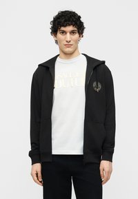 Νέος άνδρας με σκούρα σγουρά μαλλιά, ντυμένος με μαύρο φούτερ με φερμουάρ πάνω από ένα λευκό t-shirt Versace Jeans Couture, στέκεται μπροστά σε απλό φόντο.