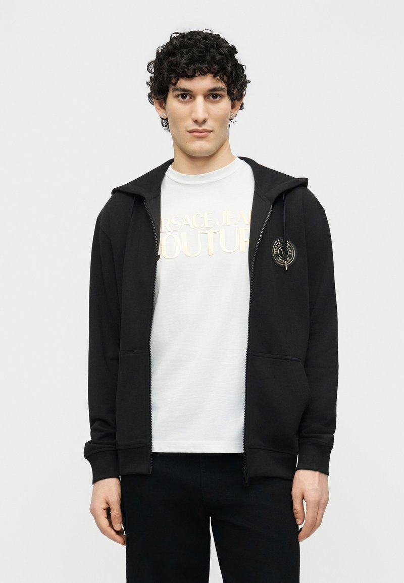 Νέος άνδρας με σκούρα σγουρά μαλλιά, ντυμένος με μαύρο φούτερ με φερμουάρ πάνω από ένα λευκό t-shirt Versace Jeans Couture, στέκεται μπροστά σε απλό φόντο.