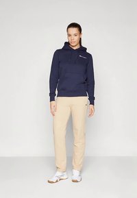 Mulher de pé com calças bege, capuz azul-marinho com logotipo, tênis brancos, contra um fundo cinza claro liso.