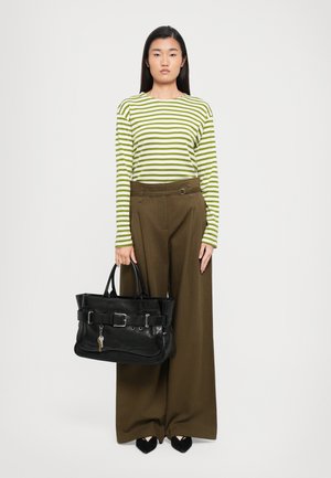 Borsa tote in pelle nera con corpo ampio, hardware in argento e dettaglio decorativo con fibbia. Il modello indossa una maglietta a righe verde e bianca e pantaloni larghi marroni.