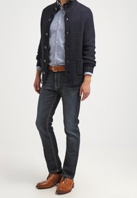 Muž v námořnickém texturovaném cardiganu přes světle modrou košili, tmavých džínách, hnědém opasku a béžových kožených formálních botách, stojící před bílým pozadím.