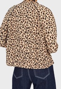 Giacca in tessuto color tan con stampa leopardata, caratterizzata da macchie nere, vestibilità rilassata, spalle arrotondate e cuciture visibili. Abbinata a jeans scuri.
