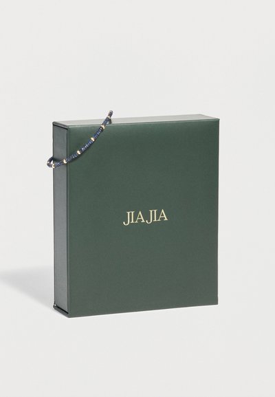Σκούρο πράσινο κουτί με χρυσά γράμματα "JIAJIA" και λεπτό μπλε και χρυσό βραχιόλι με χάντρες, τυλιγμένο μερικώς στη γωνία.
