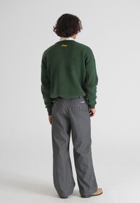 Zielony sweter z zaokrąglonym dołem i żółtym haftem; szare spodnie w paski z tylnymi kieszeniami i brązowe obuwie.