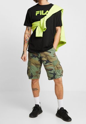Osoba nosząca czarną koszulkę FILA, spodenki cargo w kamuflażowym wzorze, białe skarpetki, czarne trampki oraz neonowozielony sweter zarzucony na ramiona.