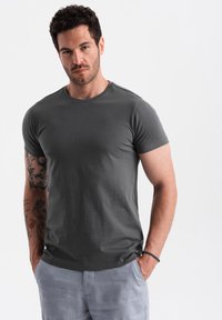 Ciemnoszary t-shirt z krótkim rękawem, okrągły dekolt, miękki bawełniany materiał, dopasowany krój, noszony z jasnoszarymi spodniami, posiada widoczne tatuaże na ramionach.