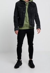 Chaqueta vaquera negra sobre una sudadera con capucha verde, combinada con jeans ajustados negros rasgados y zapatillas negras, con tejido texturizado y detalles desgastados.
