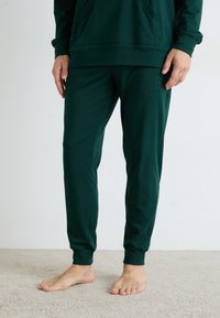 Person som har på seg mørkegrønne joggebukser og matchende hettegenser, står barføtt på lys beige teppe.