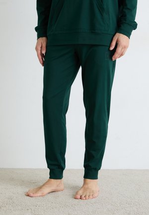 Person in einer dunkelgrünen Jogginghose und passendem Sweatshirt, barfuß auf hellbeigem Teppich stehend.