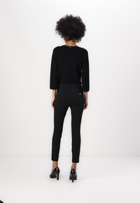 Suéter de punto negro con mangas de tres cuartos, adornado con pequeñas tachuelas en la parte posterior. Combinado con pantalones ajustados negros y tacones negros.