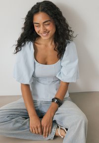 Jonge vrouw met lang, krullend haar zit in een kruiszitpositie, gekleed in een lichtblauw bovenstuk met pofmouwen, lichte denim jeans en een zwarte armband.