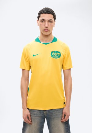 Młody mężczyzna o krótkich ciemnych włosach, ubrany w żółto-zieloną koszulkę australijskiej drużyny piłkarskiej i wyblakłe niebieskie dżinsy, stoi na tle białego tła.