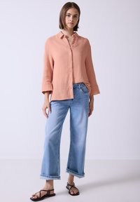 Camicia button-up rosa chiaro con vestibilità ampia e maniche arrotolate, abbinata a jeans blu a gamba larga e sandali neri con cinturini.