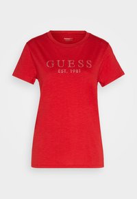 Raudonas trumpomis rankovėmis T-shirt, pagamintas iš medvilnės. Yra apvalus kaklas ir priekyje puikuojasi tekstas "GUESS EST. 1981" iš cirkonio.
