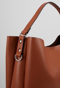 Sac cabas en cuir marron texturé avec sangle cousue et anneaux métalliques argentés ainsi que rivets sur un fond gris uni.