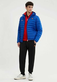 Giacca imbottita blu con cappuccio, con logo sul petto. Indossata sopra una felpa rossa, abbinata a pantaloni neri e sneakers bianche.