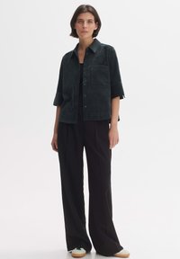 Dark green suede short-sleeve shirt with a single patch pocket, paired with high-waisted black trousers and sneakers with a light sole.