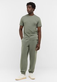 Oliwkowa koszulka z krótkim rękawem w zestawie z dopasowanymi spodniami dresowymi. Gładki materiał, luźny krój i elastyczne mankiety. Białe sneakersy dopełniają całość.