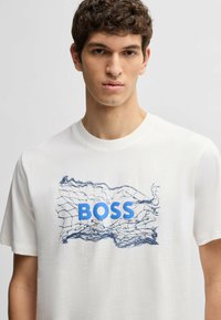 Hvid bomulds t-shirt med et blåt grafisk design, der har ordet "BOSS" og abstrakte linjemønstre. Rund hals og korte ærmer.