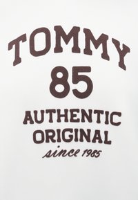 Biele bavlnené tričko s tmavo hnedým, tučným textom: "TOMMY 85 AUTHENTIC ORIGINAL since 1985." Písmo je mierne vystúpené a textúrované.
