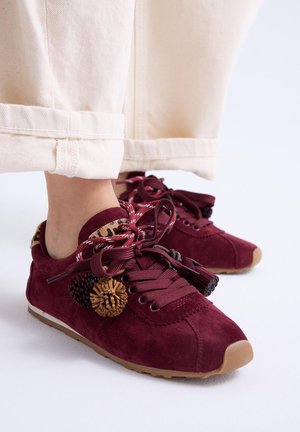 Pies con pantalones beige con puños y zapatillas burdeos con cordones gruesos y pompones decorativos sobre un fondo claro.