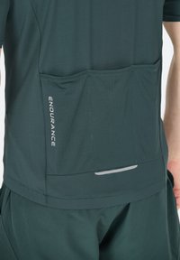 Dunkelgrünes Sportshirt mit strukturiertem Finish, ausgestattet mit einer Rückentasche und reflektierendem Streifen. Das Design enthält das Wort "ENDURANCE" in Silber.