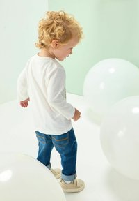 Chemise blanche à manches longues avec un design graphique, jeans en denim bleu, baskets crème ; un enfant se tient au milieu de grands ballons blancs sur un fond clair.