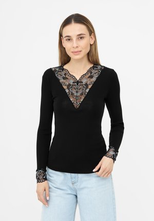 Frau trägt ein schwarzes Langarmtop mit transparenter Blumen-Spitze am tiefen V-Ausschnitt und den Bündchen, kombiniert mit hellblauen Jeans, steht vor weißem Hintergrund.