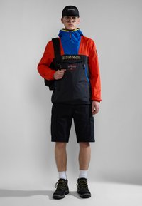 Rompevientos colorido con mangas rojas, capucha azul y cuerpo negro; presenta un parche de logo y cremalleras. Combinado con pantalones cortos cargo negros y botas.
