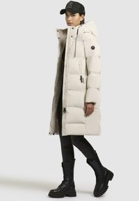 Piumino bianco con finitura testurizzata, dotato di cappuccio, tasche con zip e dettagli neri. Indossato sopra leggings neri e stivali militari.